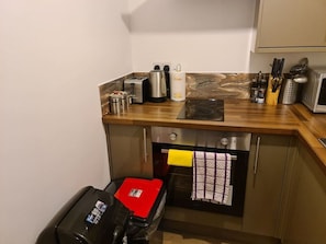 Eine kleine Küche mit Toaster, Wasserkocher und Mikrowelle auf der Arbeitsfläche. Ein roter Tablett steht auf dem Boden, und ein Mülleimer ist sichtbar.