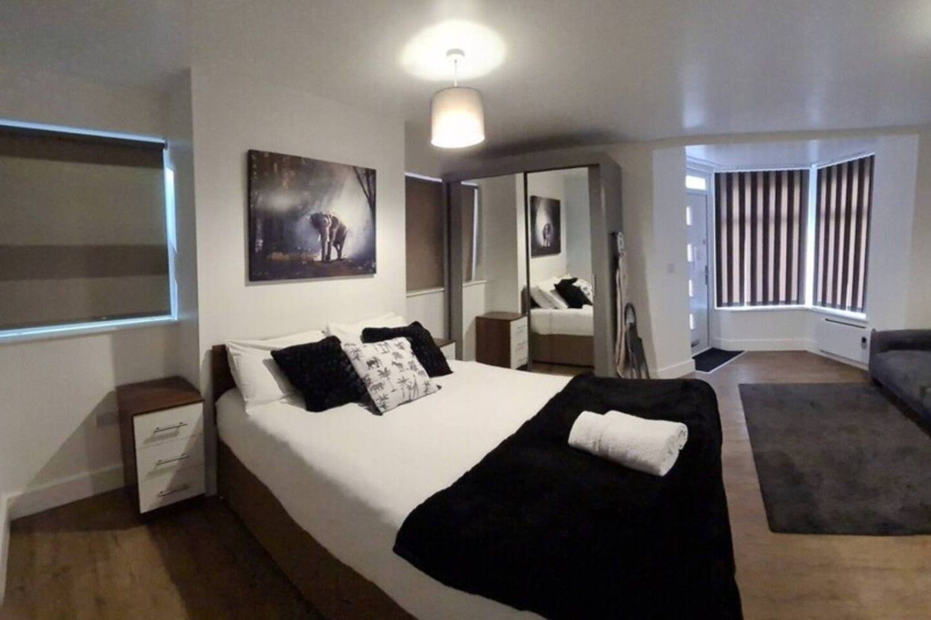 A modern hotel room with a large bed, a bedside table, a mirror, and a window with blinds.