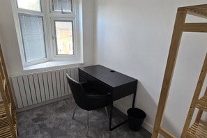 Ein kleiner, kompakter Raum mit einem einzelnen schwarzen Schreibtisch und Stuhl, einem Heizkörper, einem Fenster mit Jalousien und einer Holzleiter.