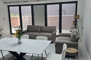 A modern living room with a sofa set, a dining table, and a large window with blinds.