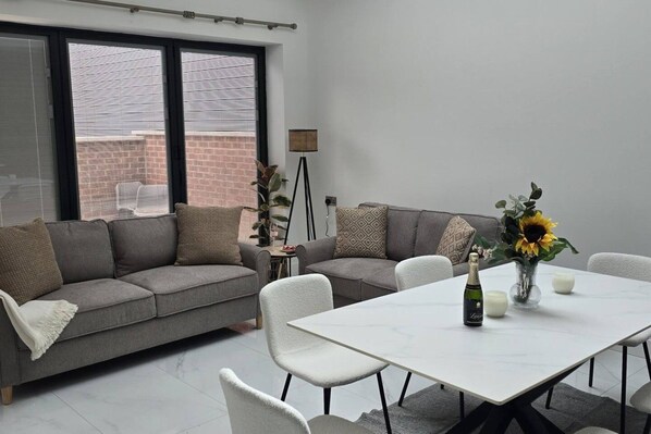 A modern living room with a grey sofa, white chairs, a dining table with a vase of sunflowers, and a floor lamp.