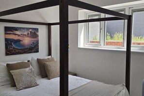 Ein Schlafzimmer mit einem Himmelbett, einem Gemälde einer Küstenlandschaft und einem Fenster mit Blick auf Grünflächen.