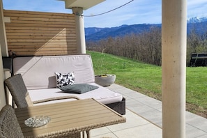 Ein überdachter Außenbereich mit einer Sitzecke, einer Couch und einem Tisch, mit Blick auf eine grüne Landschaft und Berge.