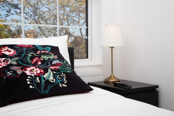 A bed with a white pillow and a black pillow with floral embroidery, a bedside lamp, and a window with a view of trees. - 4 Bed House Harrow Near Wembley (Harrow)
