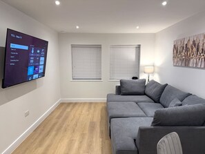 Uma sala de estar moderna com uma TV de tela plana na parede, um sofá seccional cinza, duas janelas com persianas e um piso de madeira.