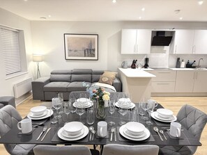 A modern dining area with a set table, grey sofas, a kitchen in the background, and a framed cityscape picture on the wall. - 2 Bed+Sofa in Sale Near Airport & Trafford Centre (Wythenshawe)
