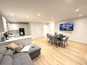 Uma sala de estar moderna com um sofá cinzento, uma mesa de jantar com cadeiras, uma área de cozinha e uma TV de tela plana na parede.