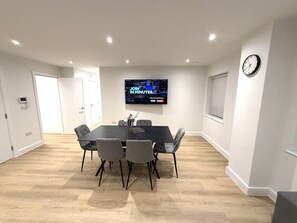 Sala de conferências com mesa preta e cadeiras cinzas, TV de tela plana na parede e relógio na parede.