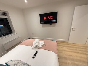 A hotel room with a bed, a TV mounted on the wall displaying the Netflix logo, a window with blinds, and a door. - 1 Bed+Sofa in Sale Near Airport & Trafford Centre (Wythenshawe)