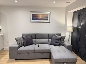 Ein modernes Wohnzimmer mit einem grauen Ecksofa, einer Fernbedienung auf der Armlehne und einem gerahmten Bild an der Wand.