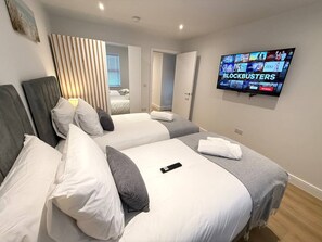 Quarto de hotel com duas camas, uma TV exibindo 'BLOCKBUSTERS' e uma janela com persianas.