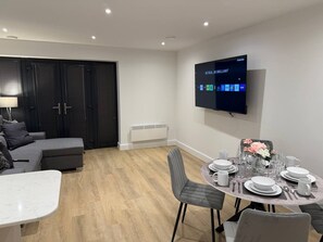 A modern living room with a grey sofa, a dining table set with plates and glasses, and a flat-screen TV mounted on the wall. - Twin+Sofa Bed in Sale Nr Airport & Trafford Centre (Wythenshawe)