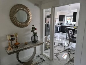 A modern interior with a marble floor, a console table with decorative items, a mirror, and a view into a kitchen area.