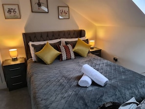 A neatly made bed with a grey quilt, multiple pillows, and two rolled towels.