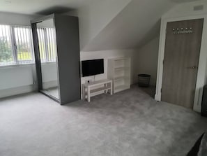 Une pièce avec un téléviseur posé sur un meuble, un banc blanc, une porte et une fenêtre avec des stores.
