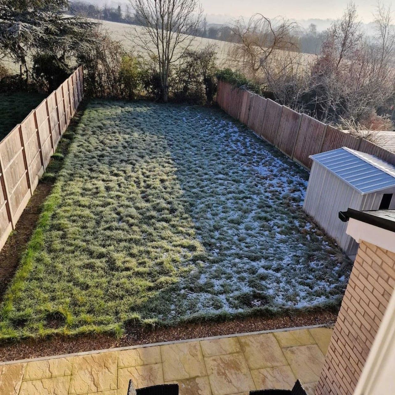 A backyard with a wooden fence, a shed, and a lawn partially covered with snow.