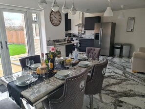 A dining area with a table set for six, a kitchen in the background, and a large clock on the wall. - SAV 5-Bed Executive Detached House, Leicester (Scraptoft)