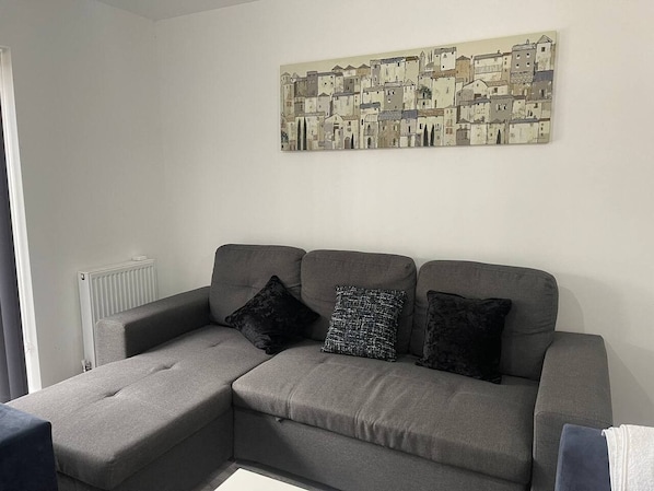 Ein graues Ecksofa mit drei schwarzen Kissen in einem Raum, der einen weißen Heizkörper und ein an der Wand befestigtes Kunstwerk zeigt.