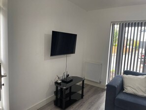 Un saló amb un televisor de pantalla plana fixat a la paret, una taula de cafè negra amb una làmpada, i un sofà blau amb un coixí blanc.