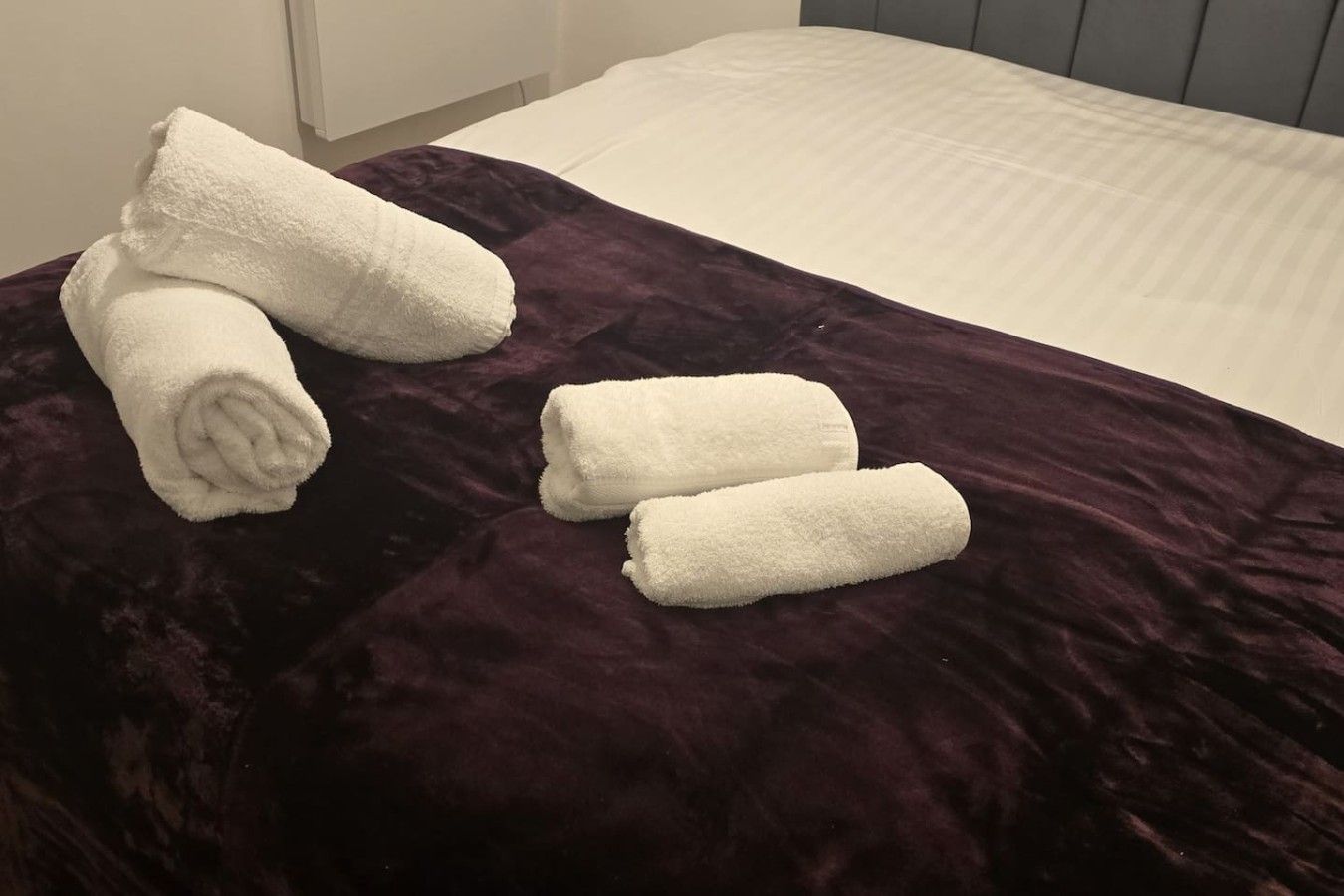 A bed with a dark purple bedspread and three rolled white towels.