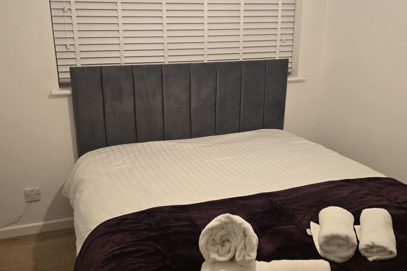 A bedroom with a bed, a headboard, a window with blinds, and a power outlet on the wall.