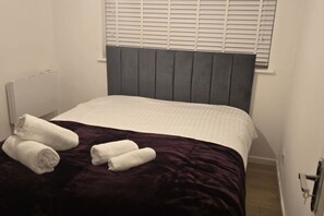 A small, single-bed room with a dark headboard, white bedding, and rolled towels on the bed. - SAV 1 Bed Flat Near Watford Town (Hertfordshire)