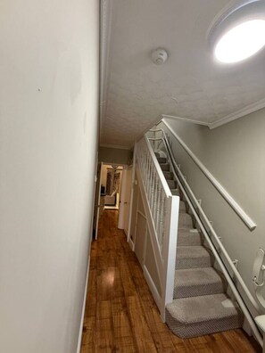 A narrow hallway with wooden flooring, a white staircase with a handrail, and a door leading to another room. - 5 Bed Family home near Heathrow l London (Greater London)