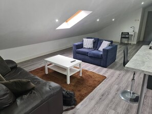 A modern loft-style living space with a high ceiling, a skylight, a blue sofa with patterned cushions, a white coffee table, and a dining area with a bar stool. - SAV Stays 2 Bed Flat, Saffron Leicester-1st Floor (Leicester)