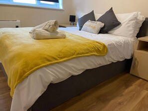 A neatly made bed with a yellow blanket and rolled towels, a bedside table with a lamp, and a wooden nightstand. - SAV 1-Bed Flat, Walking Distance to Watford Centre (Hertfordshire)