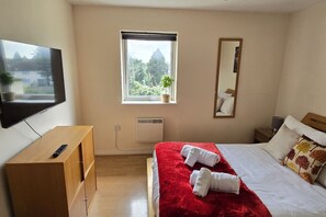 A hotel room with a bed, a TV, a window with a view of trees, a mirror, and a bedside table. - SAV 2-Bed Flat, Uxbridge (Greater London)