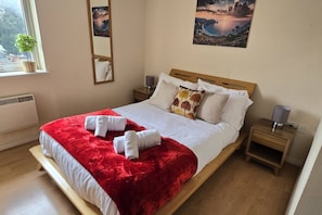 A hotel room with a bed, a red throw blanket, white towels, a nightstand with a lamp, and a window with a view of the outdoors. - SAV 2-Bed Flat, Uxbridge (Greater London)