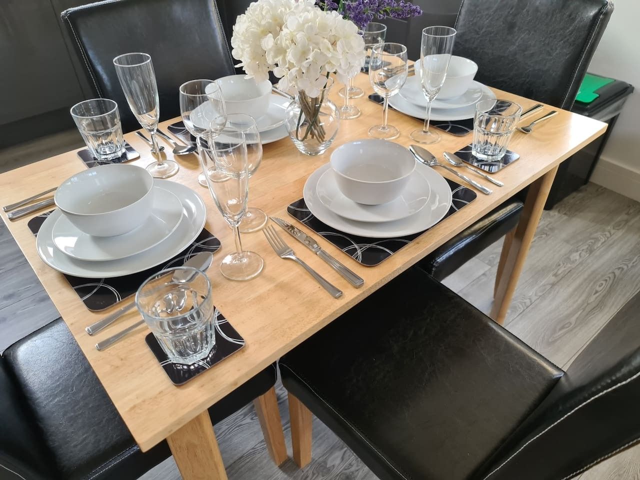 A dining table set with white plates, glasses, and cutlery, accompanied by a vase of white flowers.