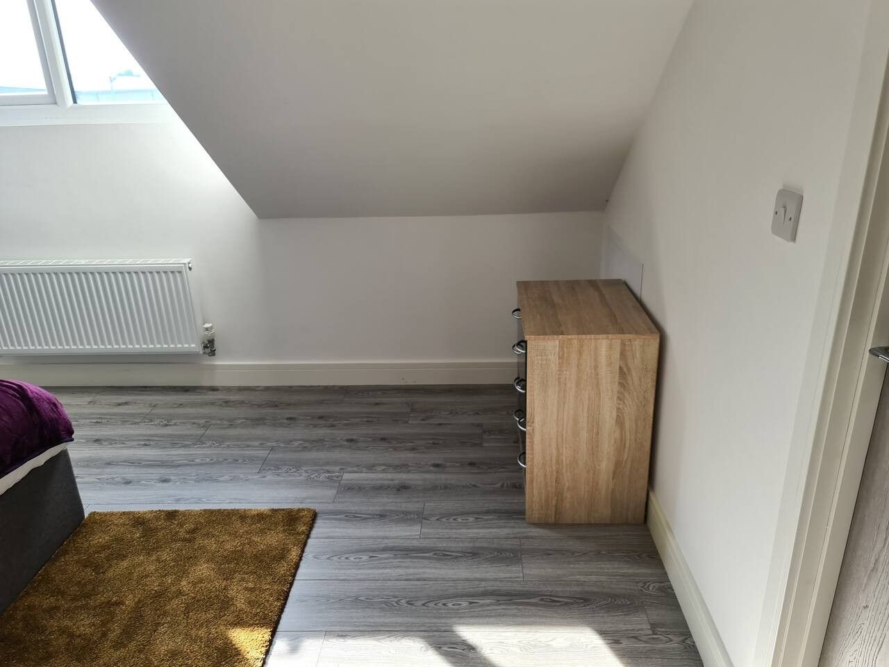 A room with a sloped ceiling, a radiator, a wooden cabinet, and a brown rug.