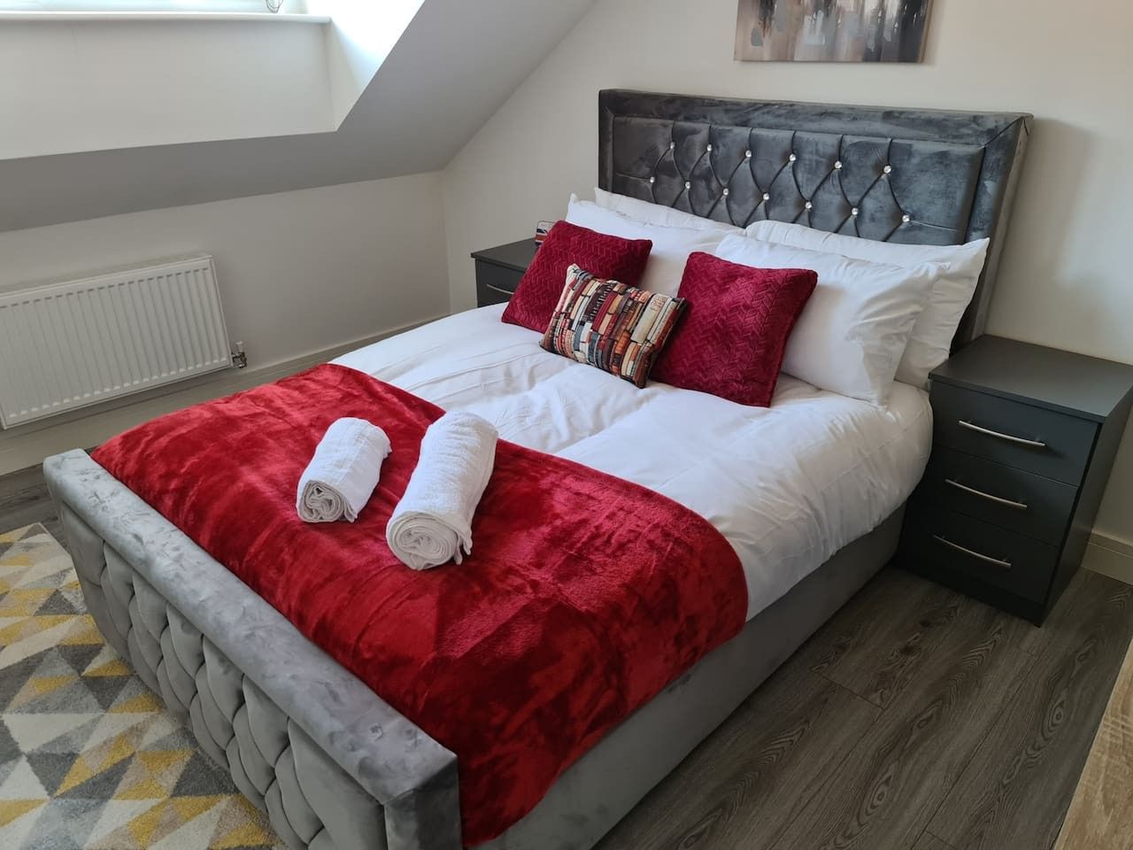 Une chambre à coucher avec un lit, deux tables de chevet, une couverture rouge et deux serviettes enroulées.