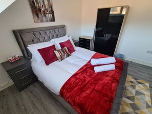 A neatly made bed with a red blanket and pillows, a bedside table with a flag, and a mirror on the wall.