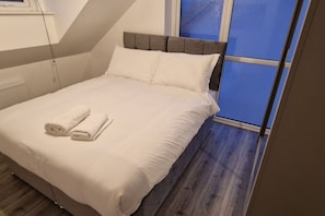 Ein ordentlich bezogenes Bett mit weißen Leinen und zwei aufgerollten Handtüchern, ein grauer möbelbezogener Kopfteil und ein Fenster mit blauen Vorhängen.