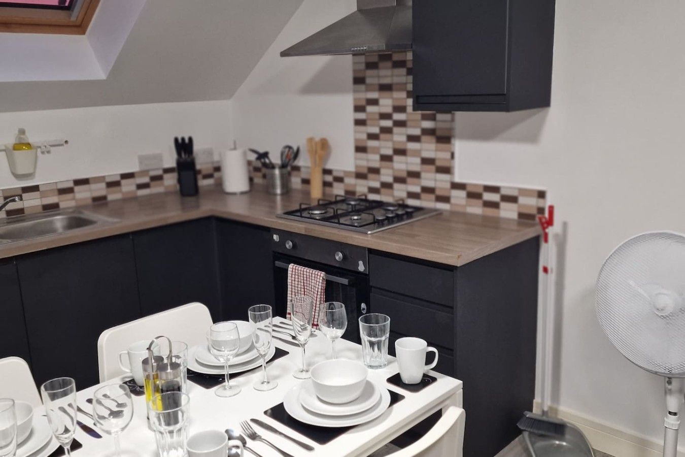 A modern kitchen with a dining area set for four, featuring a gas stove, a sink, and a fan.