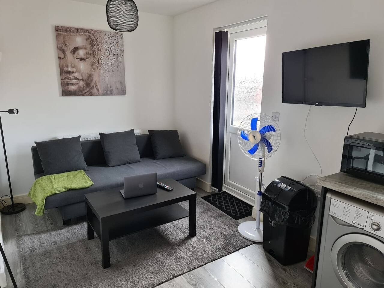 A living room with a grey sofa, a coffee table, a laptop, a wall-mounted TV, a standing fan, and a washing machine.