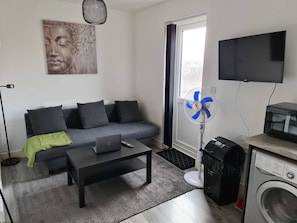 A living room with a grey sofa, a coffee table, a laptop, a wall-mounted TV, a standing fan, and a washing machine. - Sav Stays Apartments Nottingham Road - 1 Bed Flat (Leicestershire)