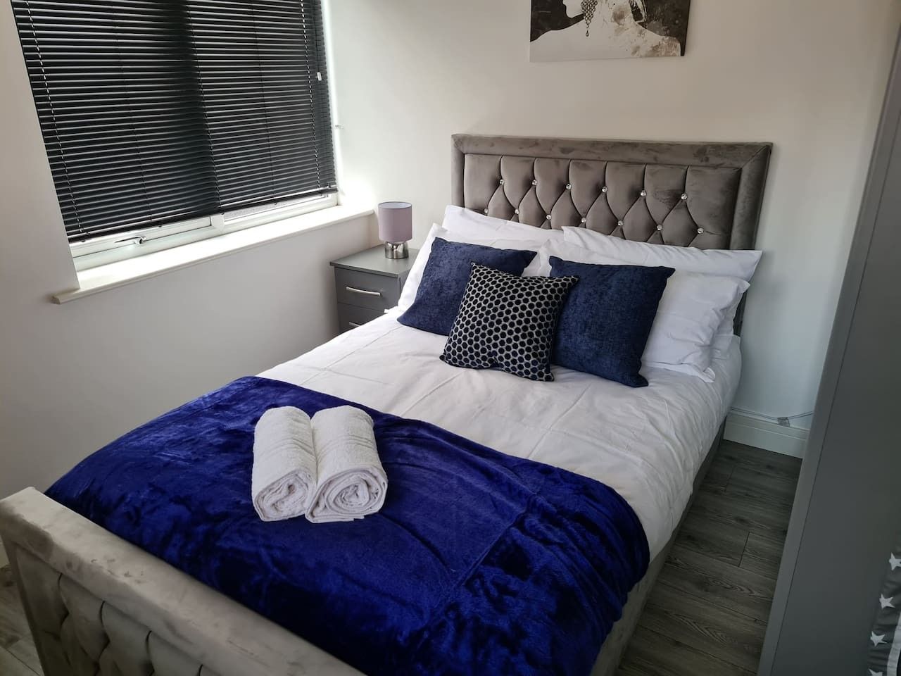 A neatly made bed with a blue blanket and rolled towels, a bedside table with a lamp, and a window with blinds.