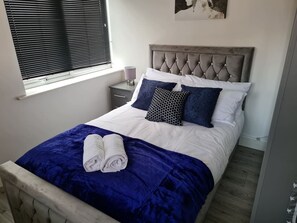 A neatly made bed with a blue blanket and rolled towels, a bedside table with a lamp, and a window with blinds. - Sav Stays Apartments Nottingham Road - 1 Bed Flat (Leicestershire)