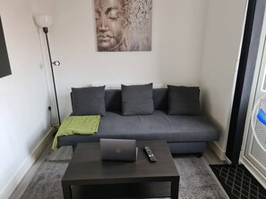 Una sala de estar moderna con un sofá gris, una mesa de centro con una computadora portátil y un control remoto, una lámpara de pie y una obra de arte colgada en la pared.