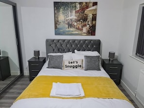 A bedroom with a tufted headboard, a yellow bedspread, and a painting of a street scene above the bed. - Sav Stays Saffron Leicester-2 Bed Flat (Leicester)