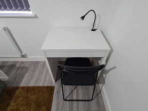 Ein weißer Schreibtisch mit einem schwarzen Stuhl, einem Heizkörper und einem Fenster mit Jalousien.