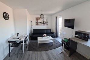 Uma sala de estar moderna com uma mesa de jantar posta para dois, um sofá preto, uma mesa de centro branca, uma TV de tela plana na parede e um micro-ondas sobre uma bancada.