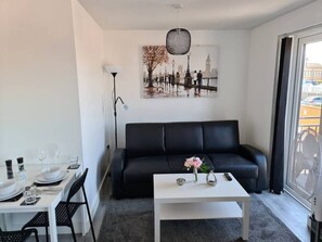 Una sala de estar moderna con un sofá negro, una mesa de centro blanca y una zona de comedor con una mesa dispuesta para dos.