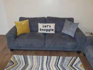 A grey sofa with two pillows, one yellow and one grey, and a cushion with the text 'Let's Snuggle'. - Sav Apartments Pretoria Watford - 1 Bedroom (Hertfordshire)