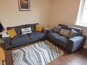 Ein Wohnzimmer mit einem grauen Ecksofa, einem gemusterten Teppich und einem kleinen Beistelltisch.