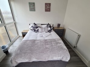 A bedroom with a bed, pillows, a nightstand, a dresser, a radiator, and a window. - Sav Apartments Nottingham Road Loughborough - 1Bed (Leicestershire)
