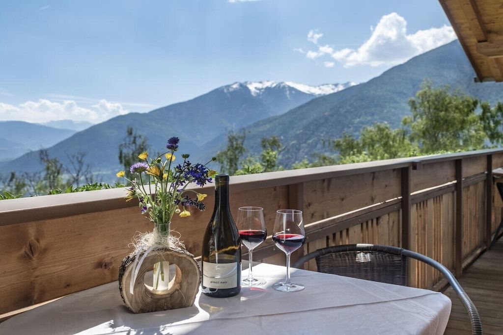 A table with a bottle of wine and two glasses on a balcony with a mountain view.